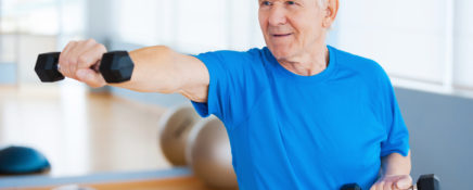 Confident senior man exercising with dumbbells and smiling while standing in health club