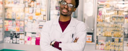 Young handsome African American man pharmacist in glasses and white coat standing in interior of modern pharmacy