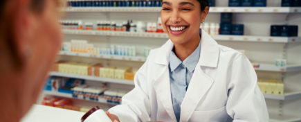 Happy female pharmacist wearing lab coat and standing behind counter giving bottle of pills to customer
