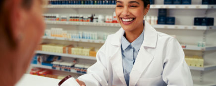 Happy female pharmacist wearing lab coat and standing behind counter giving bottle of pills to customer