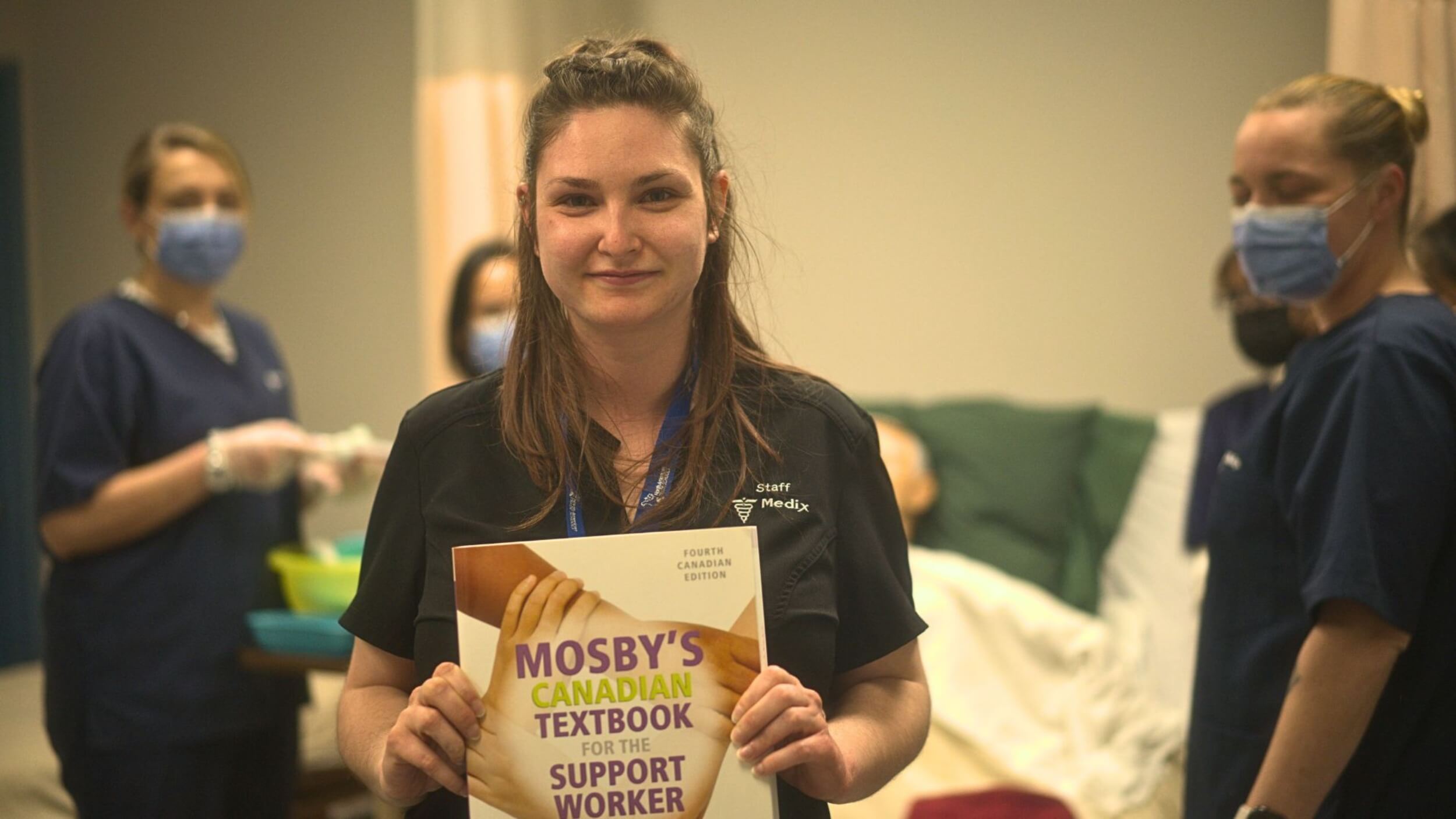 A smiling female personal support worker holding a personal support worker textbook during a practical class.