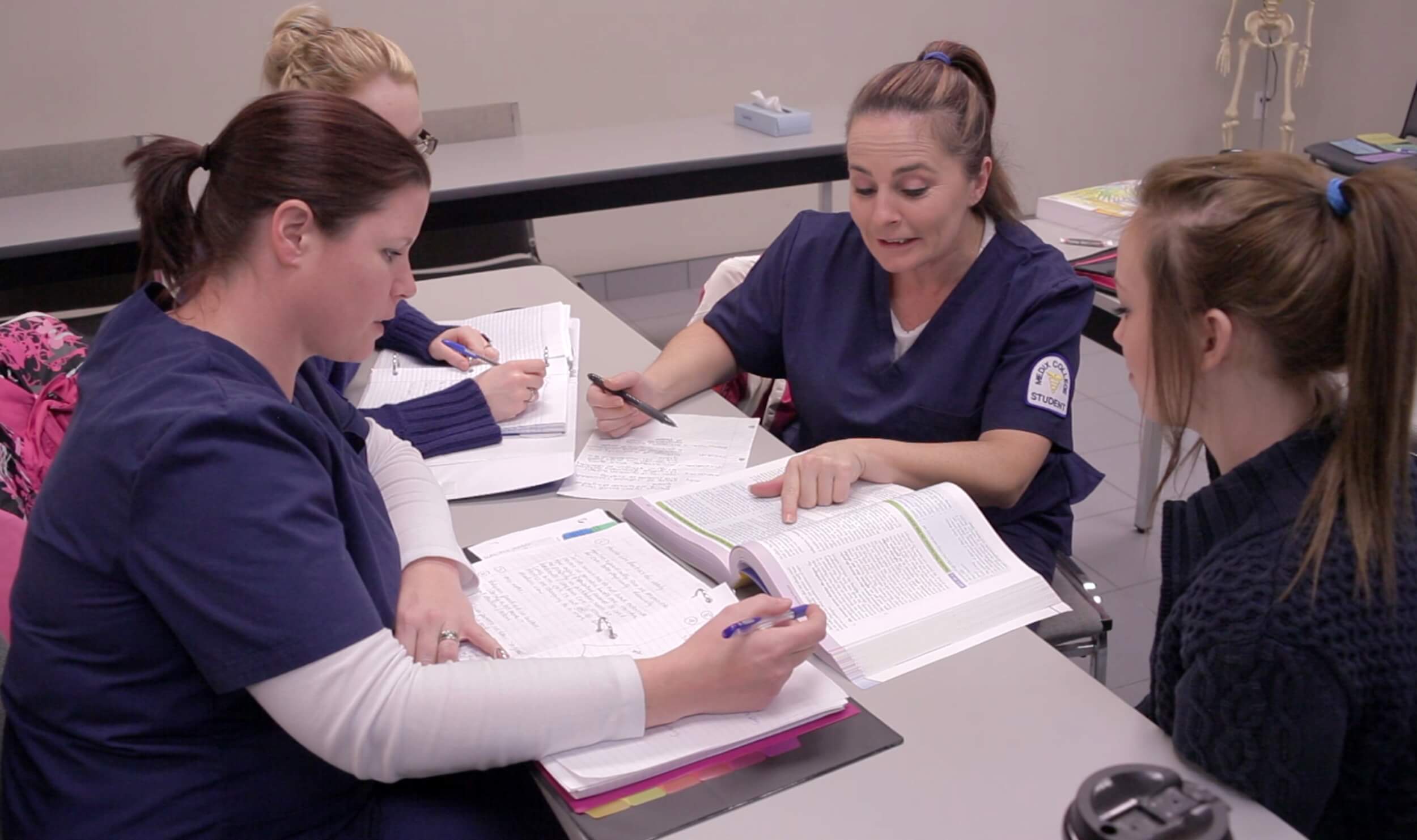 A trio of students in the community service worker course study together in a classroom, wearing navy scrubs and taking notes from textbooks.
