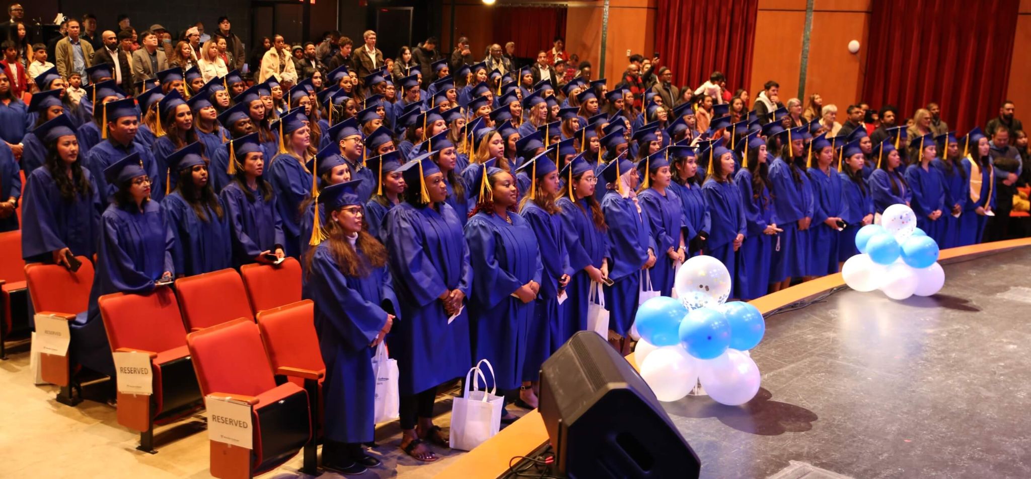 Three community service worker course graduates smile in blue graduation gowns and caps, standing outdoors with trees in the background.